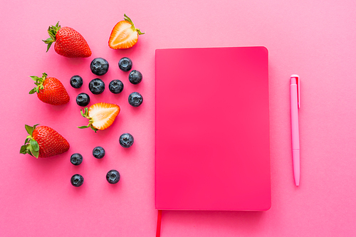 Top view of natural berries near notebook and pen on pink background