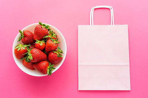 Top view of fresh strawberries in bowl and shopping bag on pink background