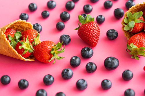 Close up view of blurred blueberries near waffle cones with strawberries on pink background