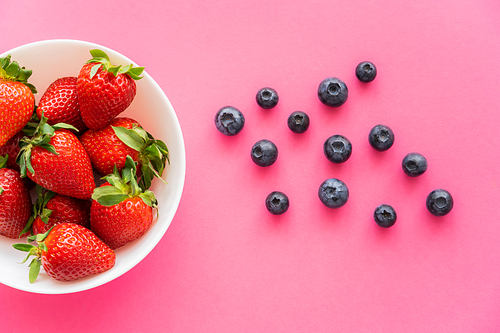 Top view of strawberries in bowl near blueberries on pink background