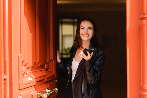 happy young woman in black leather jacket holding smartphone near orange door
