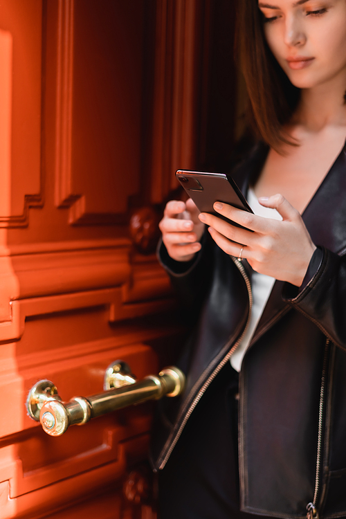young woman in black leather jacket messaging on smartphone near orange door