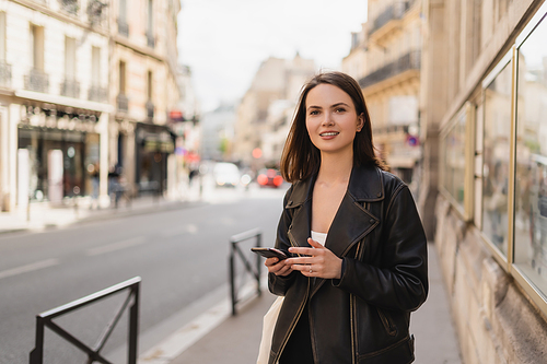 cheerful young woman in black leather jacket holding smartphone on street in paris