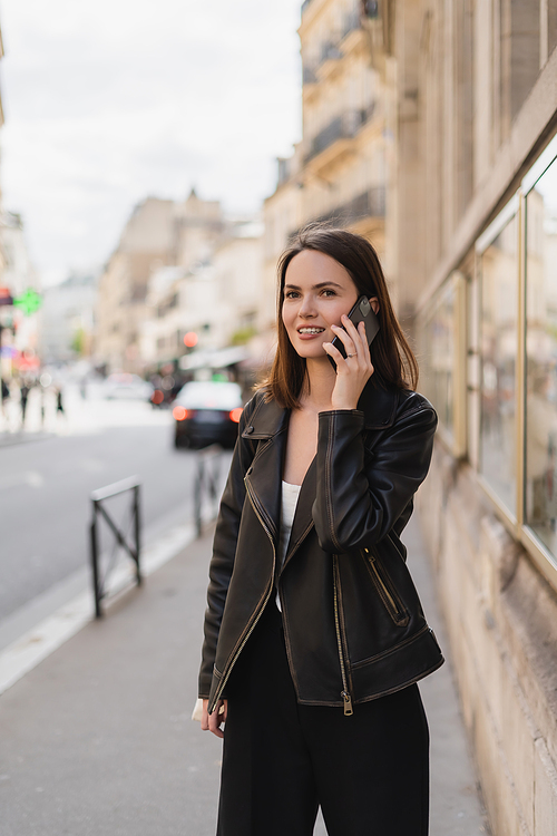 cheerful young woman in stylish jacket talking on smartphone on street in paris