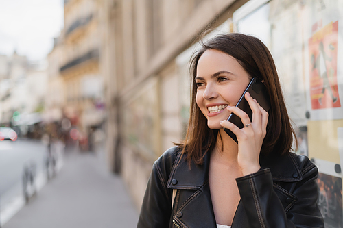portrait of joyful young woman in stylish jacket talking on smartphone on street in paris