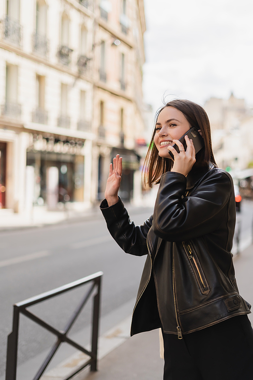 happy woman in stylish jacket talking on smartphone and waving hand on street in paris