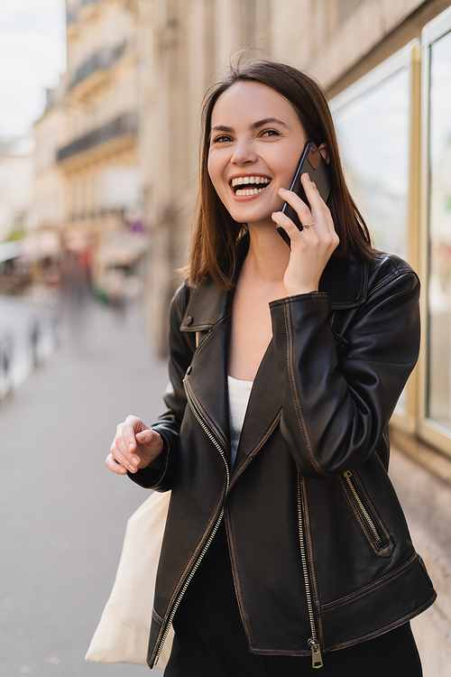 happy young woman in stylish jacket talking on smartphone and laughing on street in paris