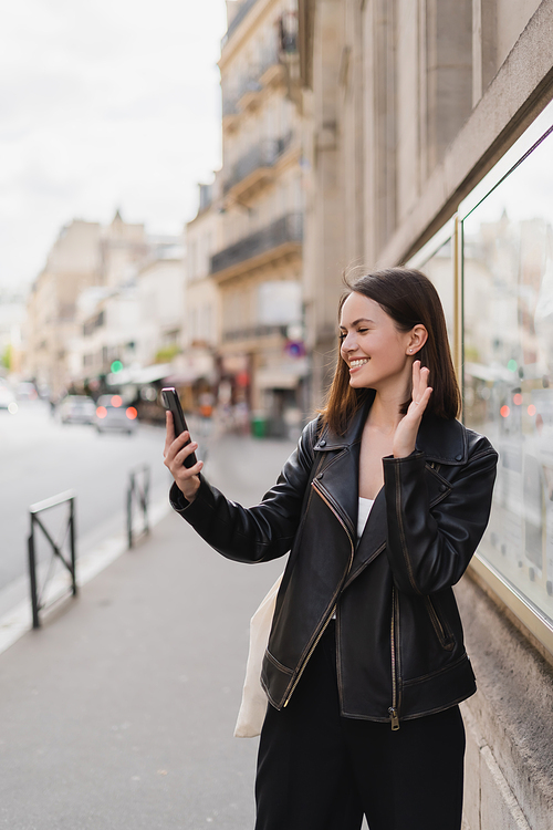 happy young woman in stylish jacket waving hand while having video chat on street in paris
