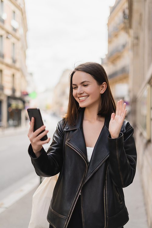 cheerful young woman in stylish jacket waving hand during video chat on street in paris