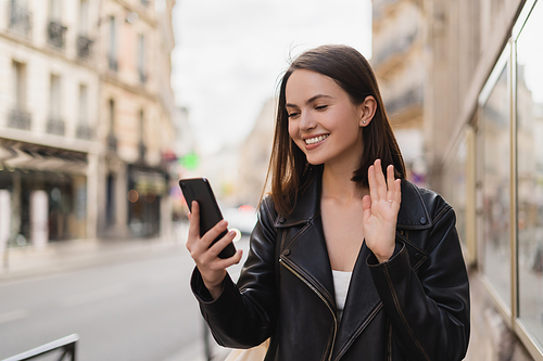 pleased young woman in stylish jacket waving hand while having video chat on street in paris
