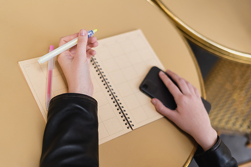 cropped view of woman holding marker pen and smartphone near blank notebook on round table