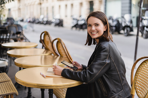 cheerful woman in leather jacket holding smartphone near notebook on round table in outdoor cafe terrace