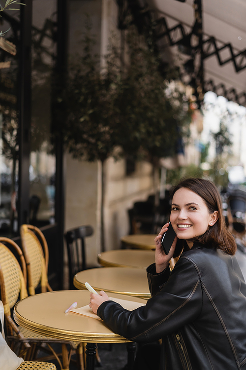 cheerful woman in leather jacket talking on smartphone and sitting in outdoor cafe in france