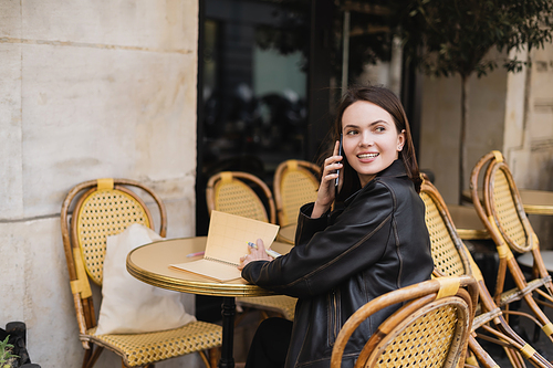 smiling woman in leather jacket talking on smartphone and sitting in outdoor cafe terrace