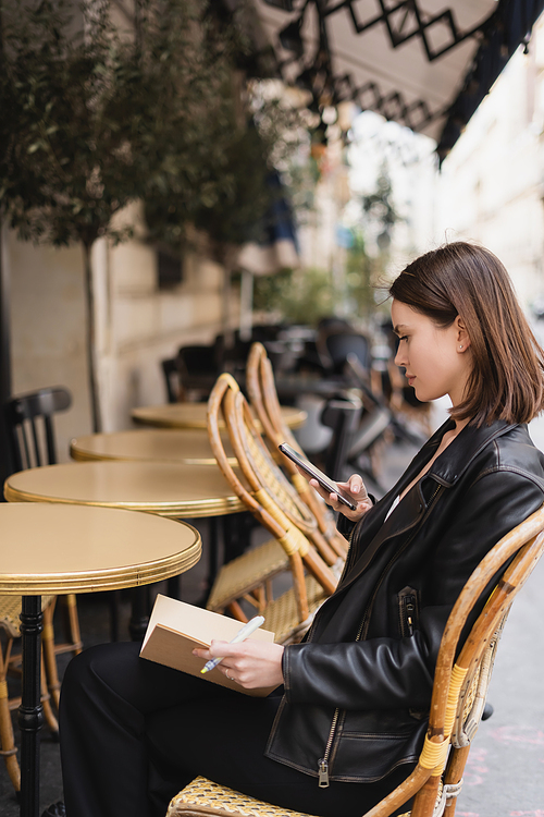 side view of woman in leather jacket holding cellphone and sitting in outdoor cafe terrace