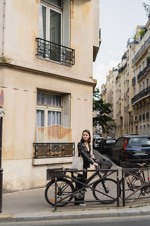 young stylish woman in black leather jacket standing near bicycle on street in paris