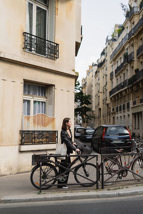 side view of young stylish woman in black leather jacket standing near bicycle on street in paris