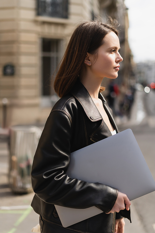 side view of young freelancer in black leather jacket walking with laptop on street in paris