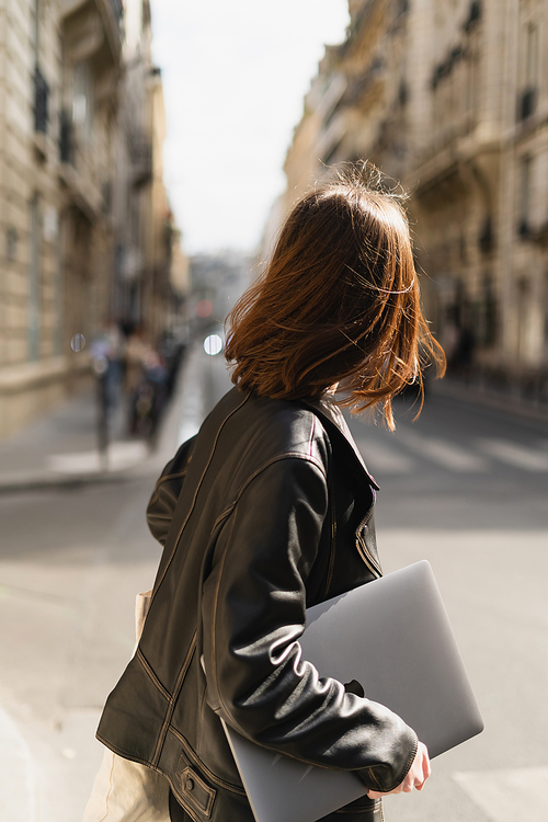 back view of freelancer in black leather jacket walking with laptop on street in paris