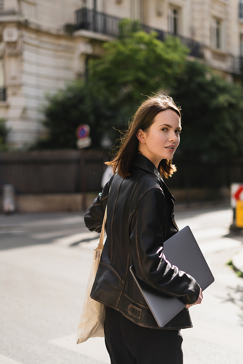 young freelancer in black leather jacket walking with laptop on street in paris