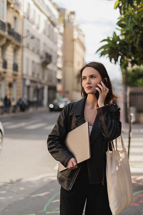young freelancer in black leather jacket holding laptop and talking on smartphone on street in paris
