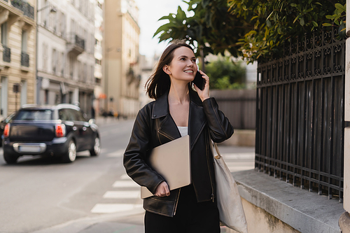 cheerful freelancer in black leather jacket holding laptop and talking on smartphone on street in paris