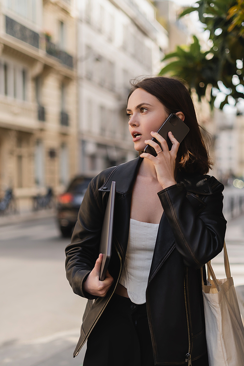 young freelancer in black leather jacket holding laptop and talking on cellphone on street in paris