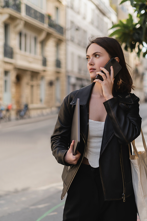 young woman in black leather jacket holding laptop and talking on smartphone on street in paris