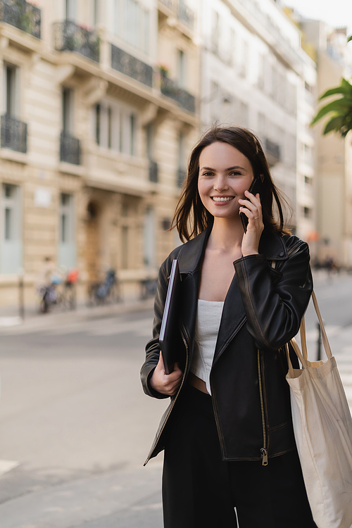 cheerful woman in black leather jacket holding laptop and talking on smartphone on street in paris