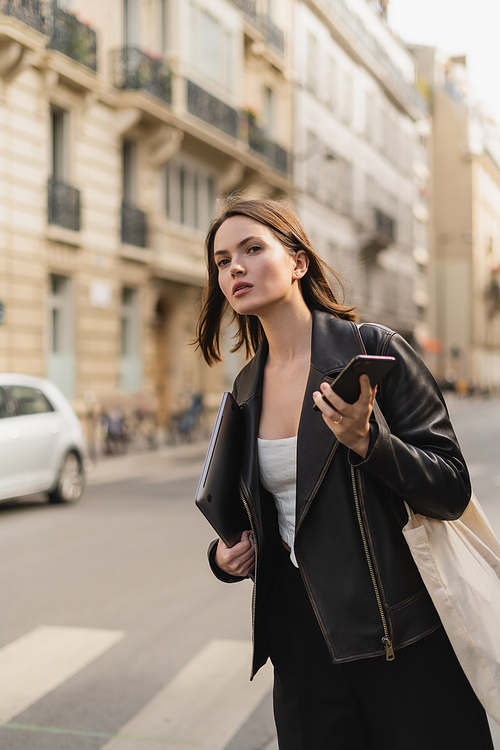 stylish woman in black leather jacket holding laptop and smartphone on street in paris
