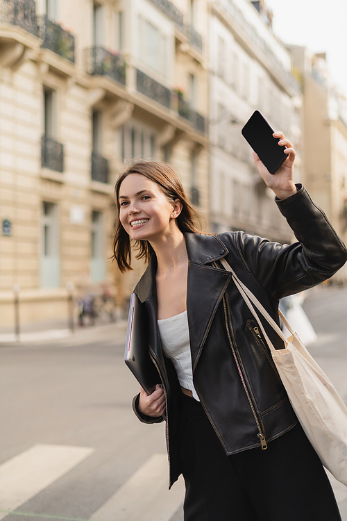 happy woman in black leather jacket holding laptop and waving with smartphone on street in paris