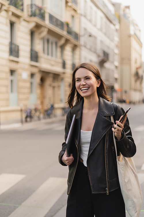 cheerful woman in black leather jacket holding laptop and smartphone on street in paris