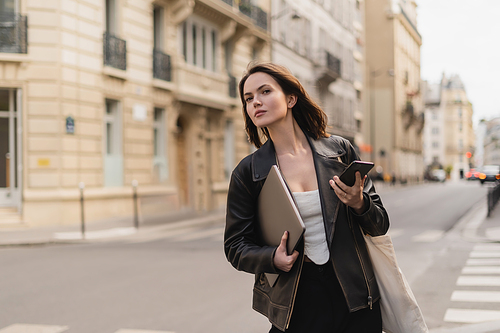 stylish freelancer in black leather jacket holding laptop and smartphone on street in paris