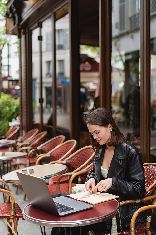 stylish freelancer in black leather jacket looking at notebook while sitting near laptop in french outdoor cafe