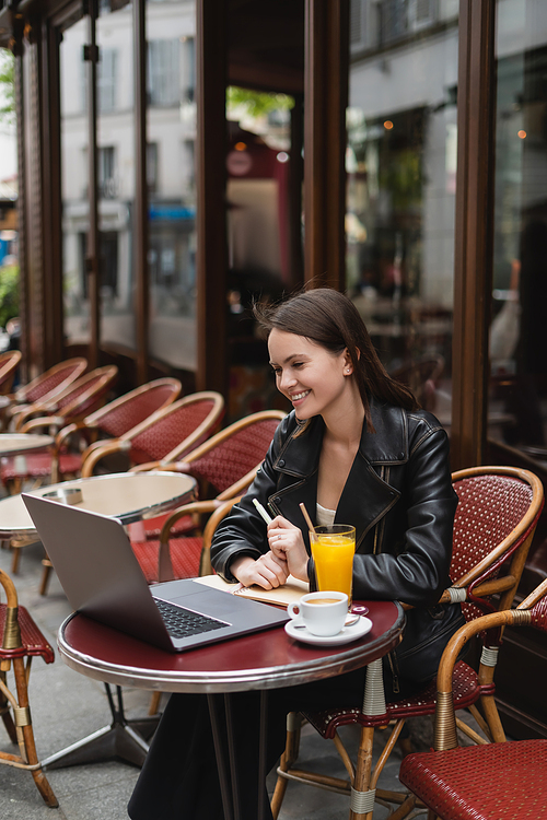 happy freelancer in black leather jacket looking at laptop near drinks on table in french outdoor cafe