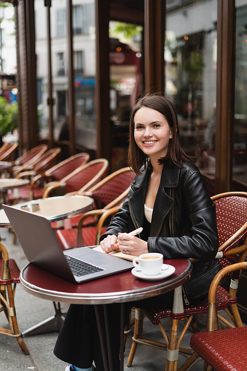 happy freelancer in black jacket  near laptop and cup of coffee on table in french outdoor cafe