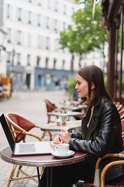 side view of smiling freelancer in black jacket looking at laptop near cup of coffee on table in french outdoor cafe