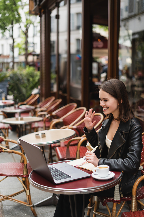 happy freelancer in black jacket having video call near cup of coffee on table in french outdoor cafe