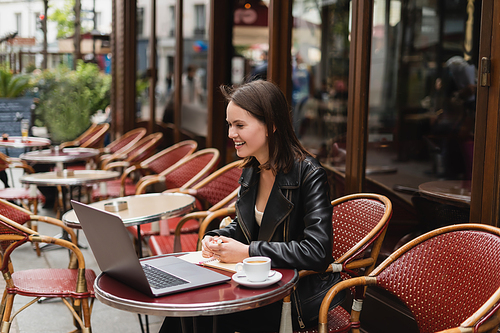 smiling freelancer in black jacket looking at laptop near cup of coffee on table in french outdoor cafe