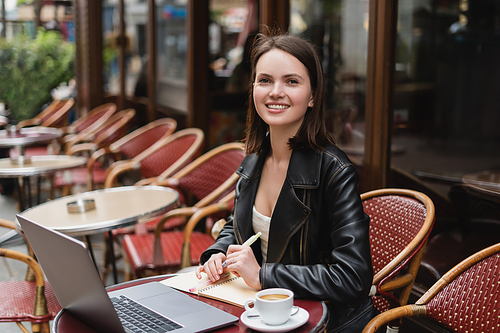 happy woman in black jacket  near laptop and cup of coffee on table in french outdoor cafe