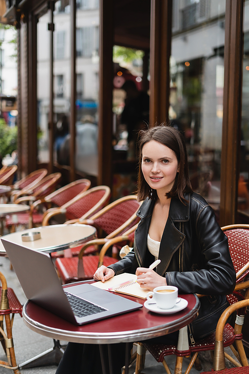 cheerful woman in black jacket  near laptop and cup of coffee on table in french outdoor cafe
