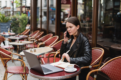 young woman in black jacket talking on smartphone near laptop and cup of coffee on table in french outdoor cafe