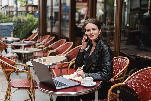 smiling freelancer in black jacket talking on smartphone near laptop and cup of coffee on table in french outdoor cafe