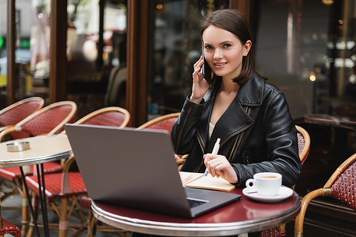 smiling freelancer in black jacket talking on smartphone near laptop and cup of coffee on table in outdoor cafe in paris