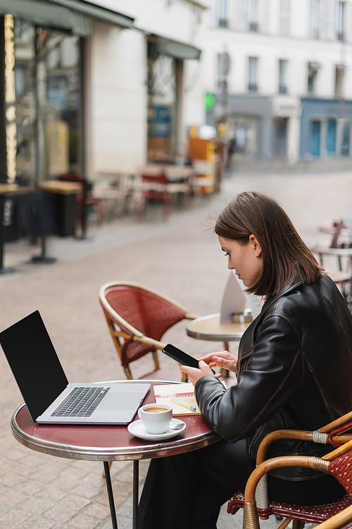 side view of freelancer in black jacket holding smartphone near laptop and cup of coffee in french outdoor cafe