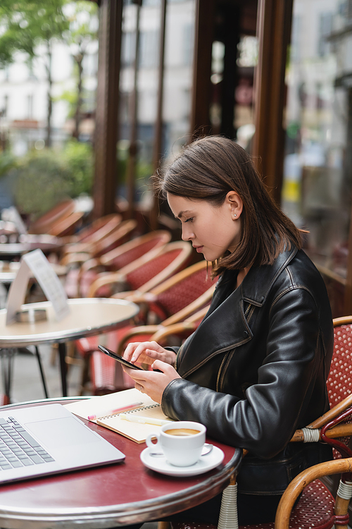 side view of freelancer in black jacket using smartphone near laptop and cup of coffee in french outdoor cafe