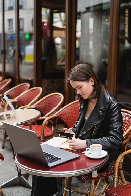 young freelancer in black jacket using smartphone with blank screen near laptop and cup of coffee in french outdoor cafe