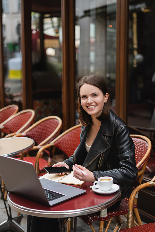 happy freelancer in black jacket holding smartphone near laptop and cup of coffee in french outdoor cafe
