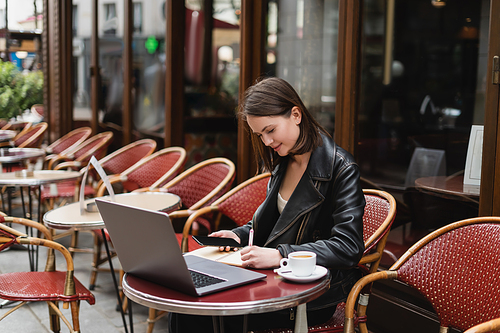young freelancer in black jacket holding smartphone and writing near laptop and cup of coffee in french outdoor cafe