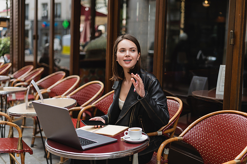 young freelancer in black jacket holding smartphone and waving hand near laptop and cup of coffee in french outdoor cafe